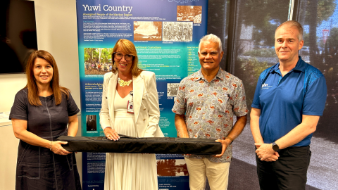 Northern Queensland PHN CEO Sean Rooney (right), Executive Director Health Service Integration and Innovation Karin Barron (left), and Traditional Custodian Elder for the Mackay region Philip Kemp present Mackay HHS CEO Susan Gannon with an updated 2024 Yuwi banner. 
